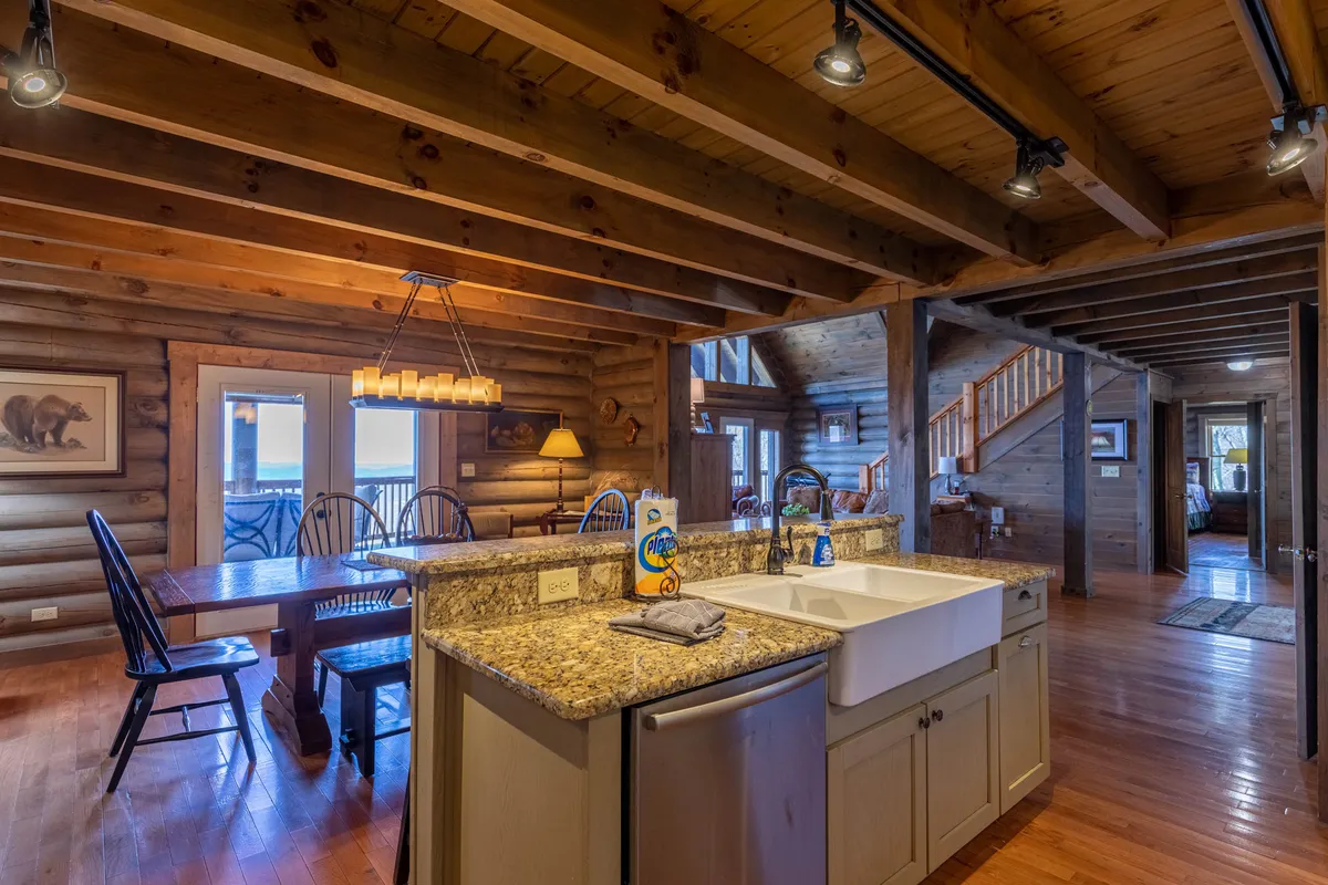 This image focuses on the dining experience, with a granite countertop leading to a large wooden dining table set beneath exposed beams and near expansive windows. The room is filled with warm light, and the inviting setup is ideal for family meals and celebrations.