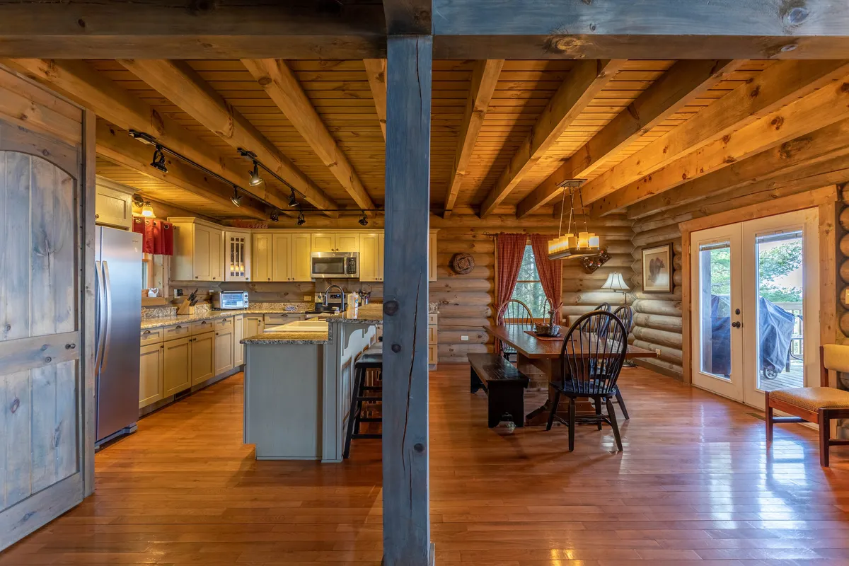 The image presents an open-concept kitchen and dining area with a prominent kitchen island, a substantial wooden dining table, and large windows that frame scenic views. Wooden beams overhead add rustic charm, while modern kitchen appliances are visible, suggesting a space designed for both cooking and gathering.