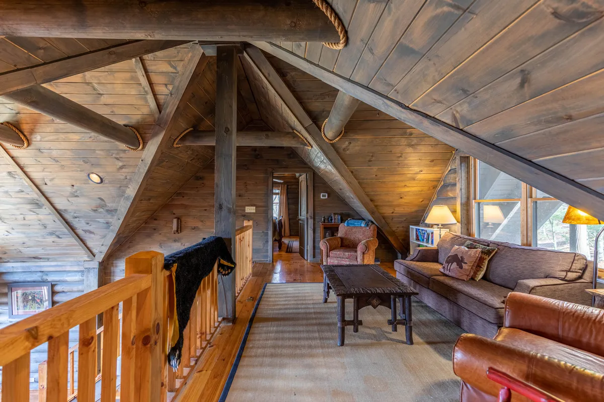 The photo features a welcoming seating area with a sofa, armchairs, a coffee table, and striking wooden beams. Large windows frame the lush landscape, offering abundant natural light and a seamless visual connection to the outdoors.