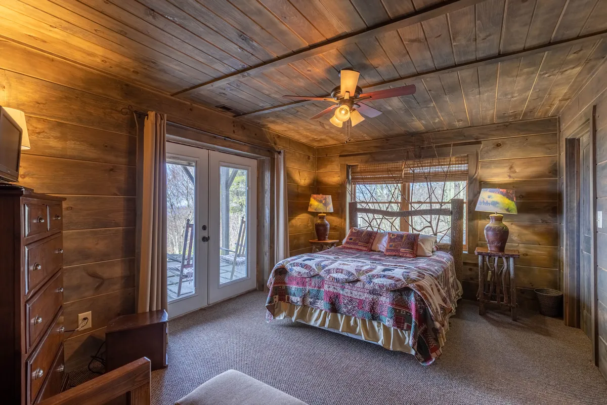 A warmly-lit bedroom featuring a bed with a patterned quilt, rustic wooden furniture, and French doors that open to beautiful nature views outside. The inviting atmosphere highlights space for relaxation and quiet moments, ideal for restful nights or reading by the window.