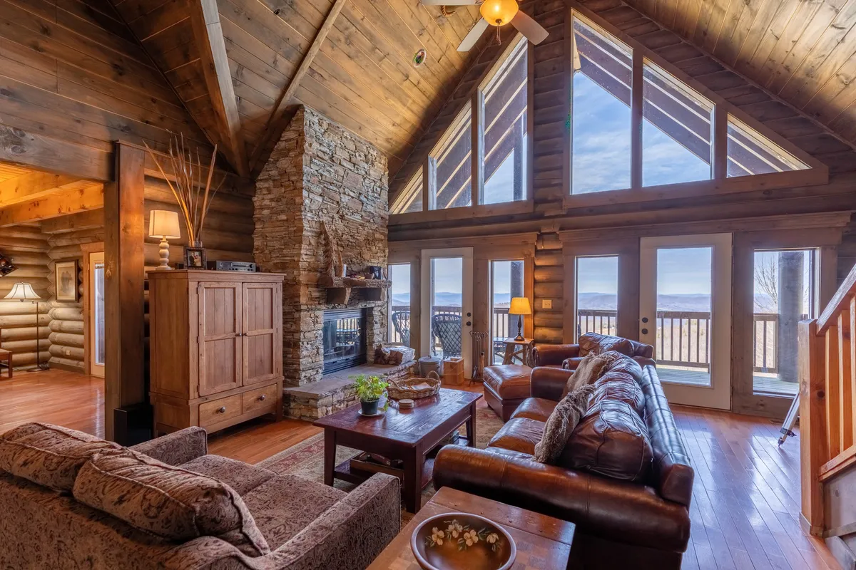 This image showcases a spacious living area with a stunning stone fireplace, large windows that frame scenic views, exposed wooden beams, and plush, comfortable seating. The natural light pouring in emphasizes the blend of rustic textures and contemporary elegance, giving a sense of connection to the outdoors.