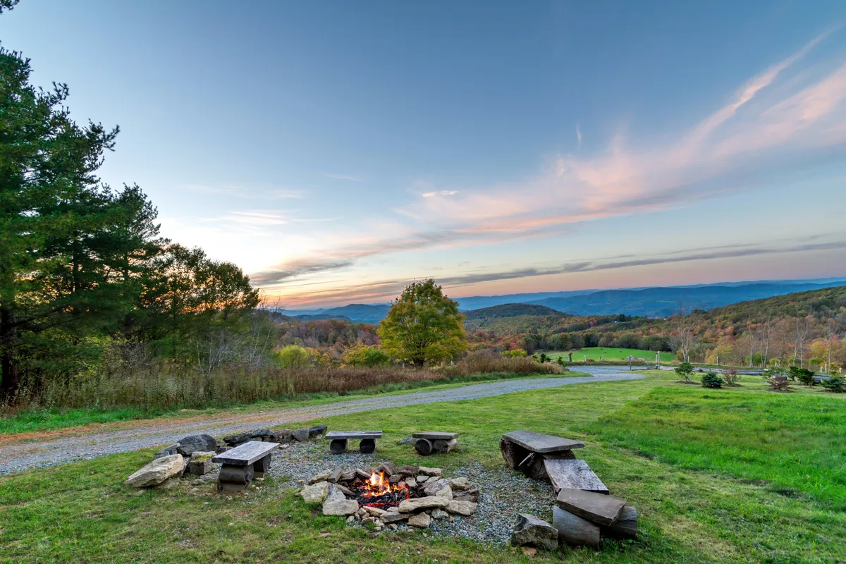 A cozy exterior fire pit scene with benches around a stone pit, distant mountain views, and a glowing sunset sky. The setup invites guests to gather for s’mores, stories, and stargazing under Banner Elk’s night sky.