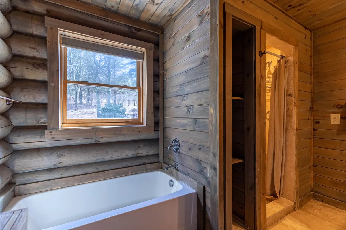 A tranquil bathroom featuring a bathtub and separate shower area, with warm wooden walls and a window offering natural light and forest views. The space evokes a spa-like feel, promising relaxation and rejuvenation after a day in the mountains.