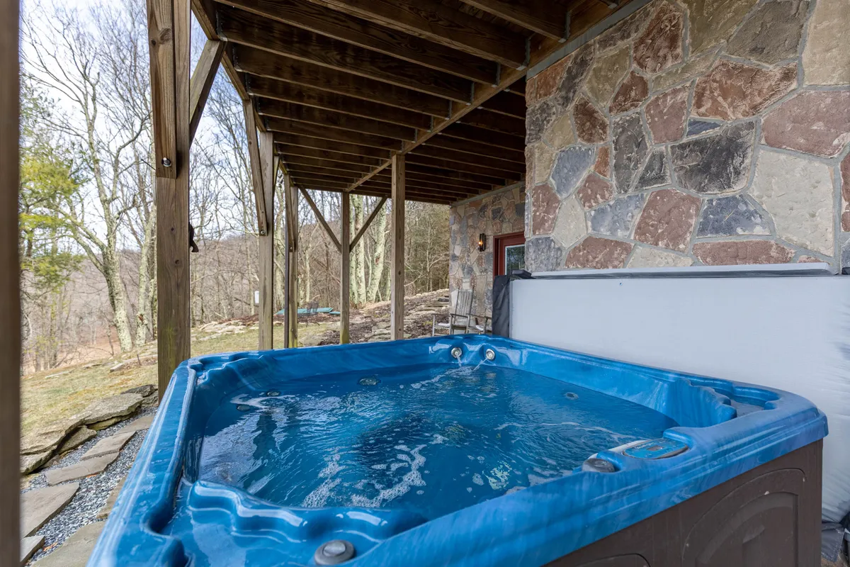 An exterior shot showcasing a hot tub set against a stone wall with exposed wooden beams, surrounded by lush natural landscaping. The spacious area is perfect for relaxing after a day spent exploring hiking trails with your dog.