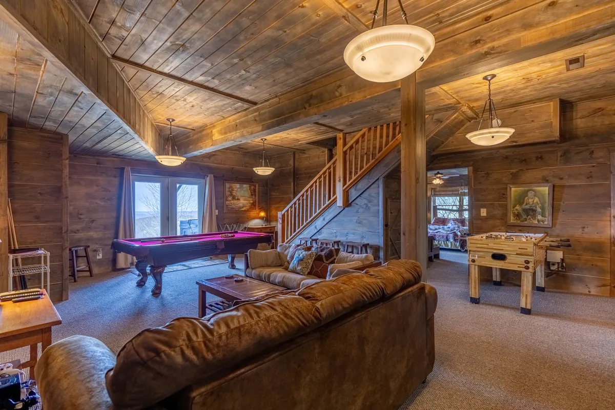 A spacious game room featuring a pool table, foosball table, inviting sofa, staircase, and large windows, all set against warm wooden walls. The setup exudes a lively, welcoming atmosphere perfect for games, relaxation, and social gatherings.