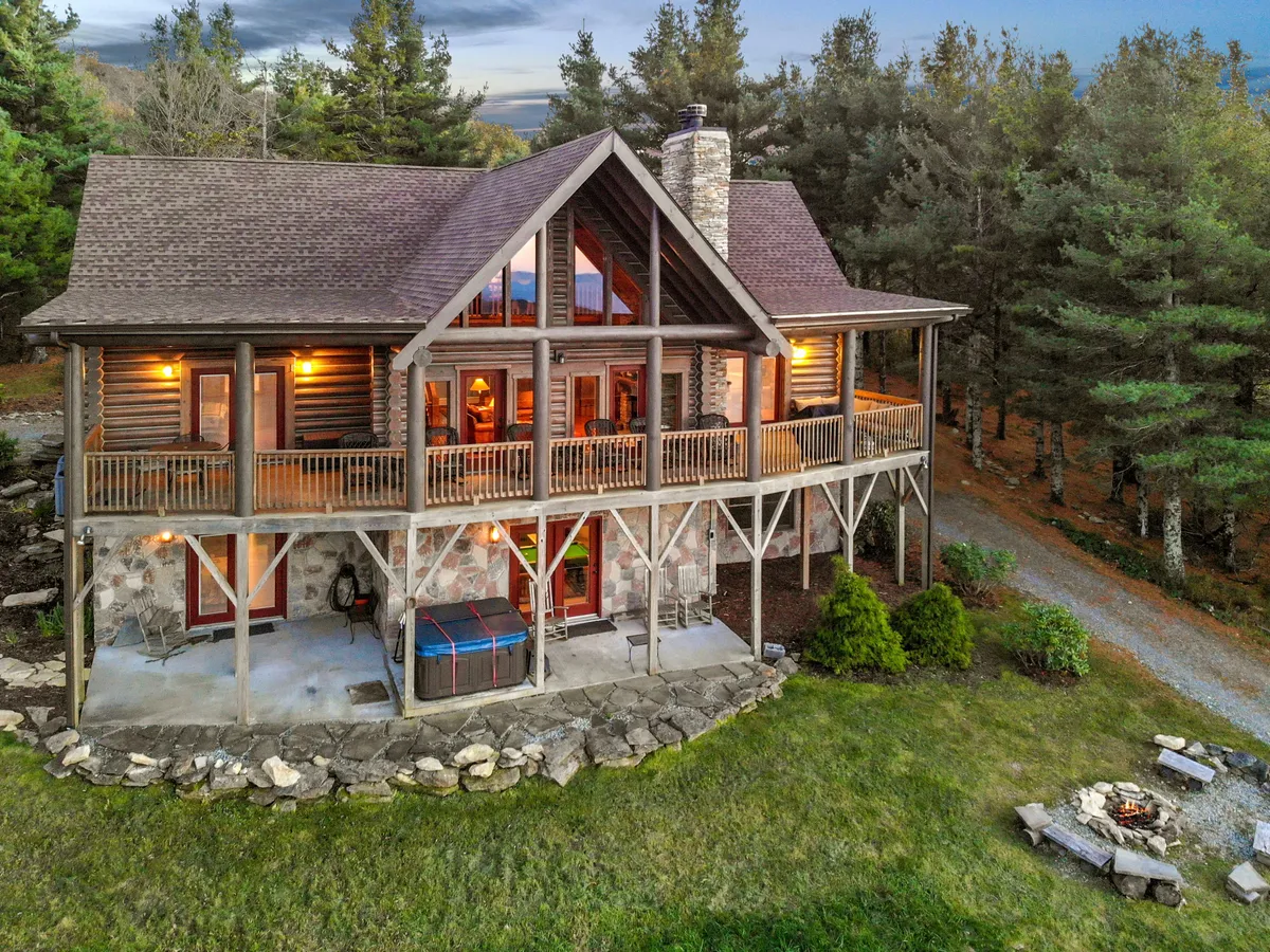 The image showcases a charming log cabin nestled in the mountains, featuring expansive windows, a spacious balcony overlooking stunning vistas, a bubbling outdoor hot tub, and a crackling fire pit. The tranquil setting is surrounded by natural beauty, inviting relaxation and connection.