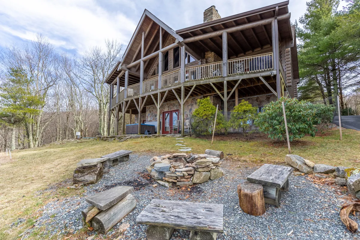 A spacious wooden deck surrounded by trees, featuring a large stone fire pit with ample bench seating around it and a hot tub in the corner. The deck offers plenty of space for outdoor gatherings, relaxation, and enjoying the natural setting.