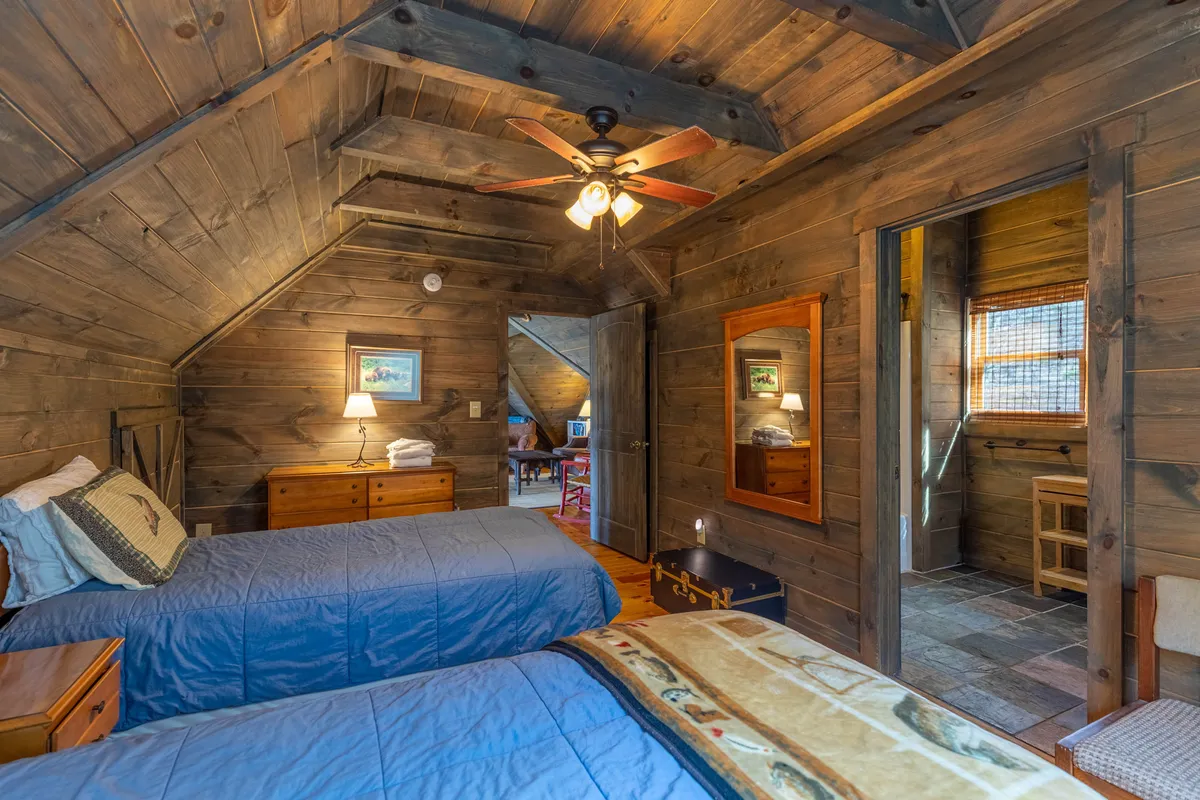A cozy upstairs bedroom with two twin beds under a wooden ceiling, complemented by a lamp, mirror, and door leading to another room. Soft natural light streams in, making the room feel peaceful and private, perfect for families or friends seeking comfort and solitude.
