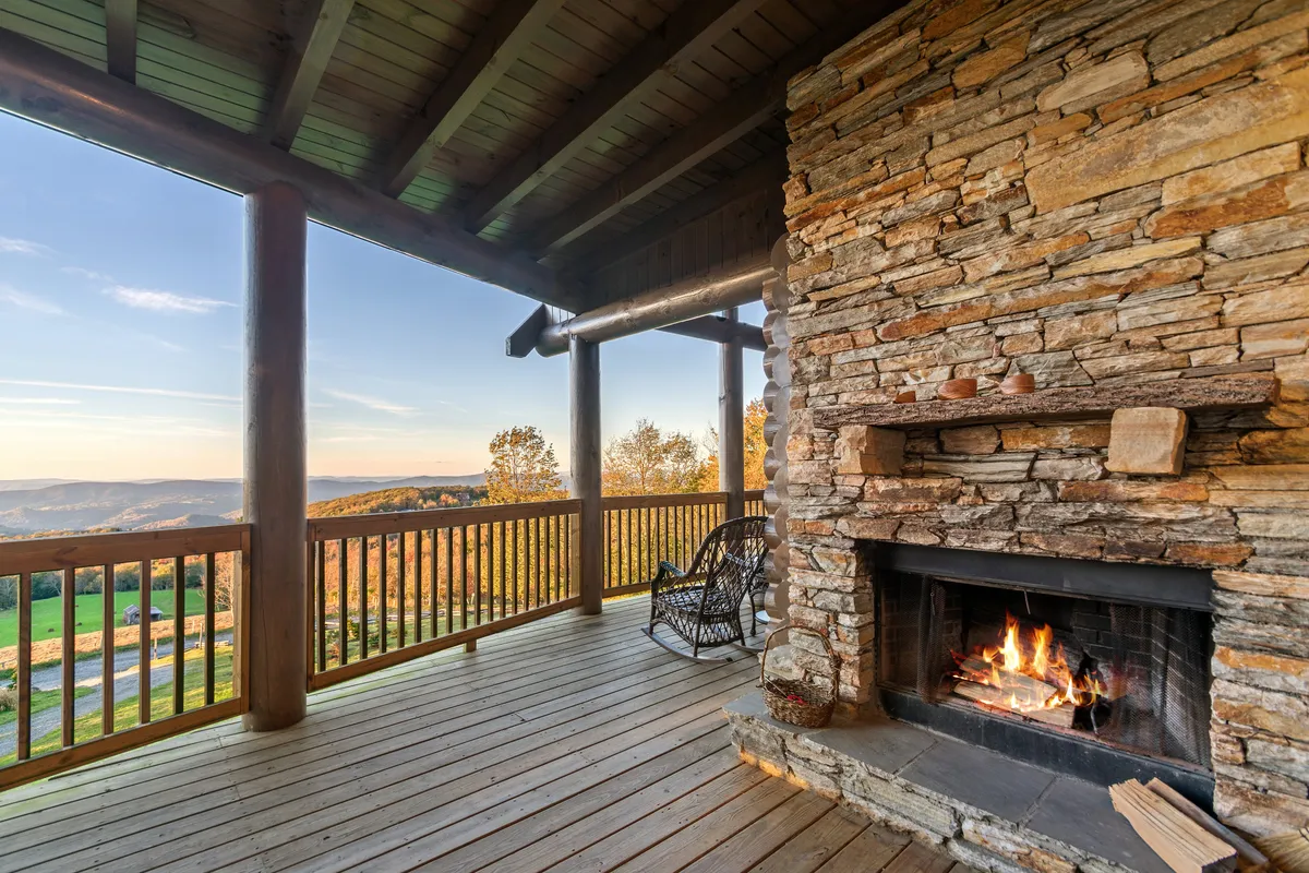 An inviting deck corner with a stone outdoor fireplace, a classic rocking chair, and panoramic mountain vistas. The scene evokes tranquility and warmth, perfect for sunset contemplation or sharing stories by the fire.