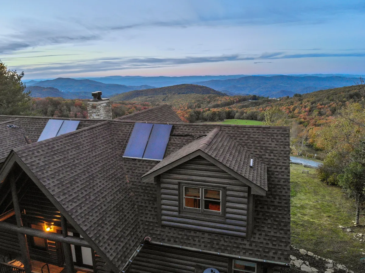 A wide-angle view of the log cabin nestled in the mountains, with solar panels on the roof and a backdrop of trees in vibrant autumn colors. The mountain landscape stretches out behind the cabin, providing a sense of the area's natural beauty and tranquility.