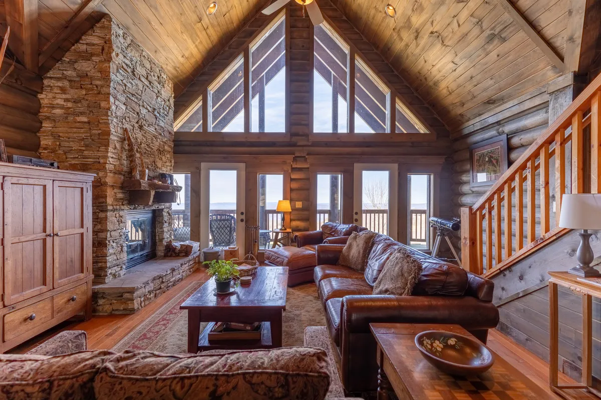 This image showcases the grand great room, featuring a large stone fireplace as the focal point. Vaulted wooden ceilings with exposed beams soar overhead, while expansive windows flood the space with natural light and offer mountain views. Comfortable leather sofas are arranged for conversation or relaxation by the fire, and a wooden staircase adds architectural interest.