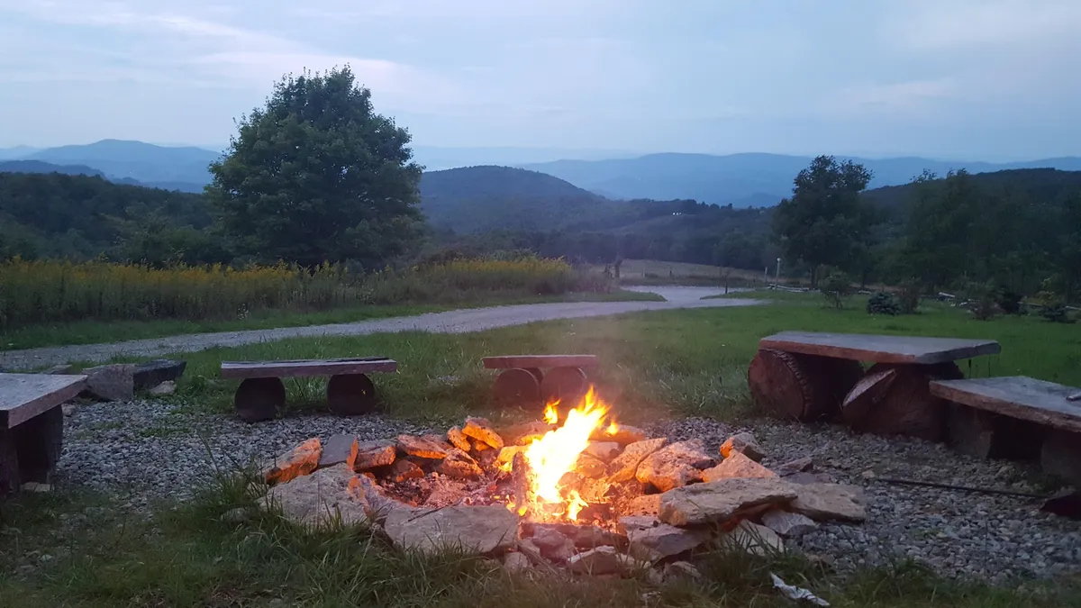 A cozy fire pit area with surrounding benches, set against the backdrop of distant mountains and nestled among trees. The setting evokes a sense of togetherness, perfect for storytelling or relaxing evenings under the stars.