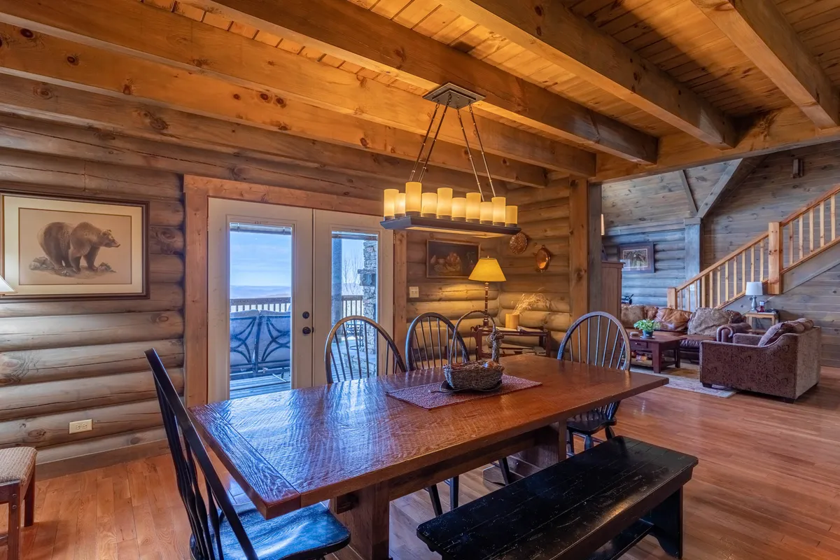 A large wooden dining table sits beneath a statement chandelier, with glass doors offering a direct view of the deck and the landscape beyond. The scene includes a comfortable seating area and mountain artwork, reinforcing a sense of place and inviting gatherings that blend indoor comfort with outdoor scenery.